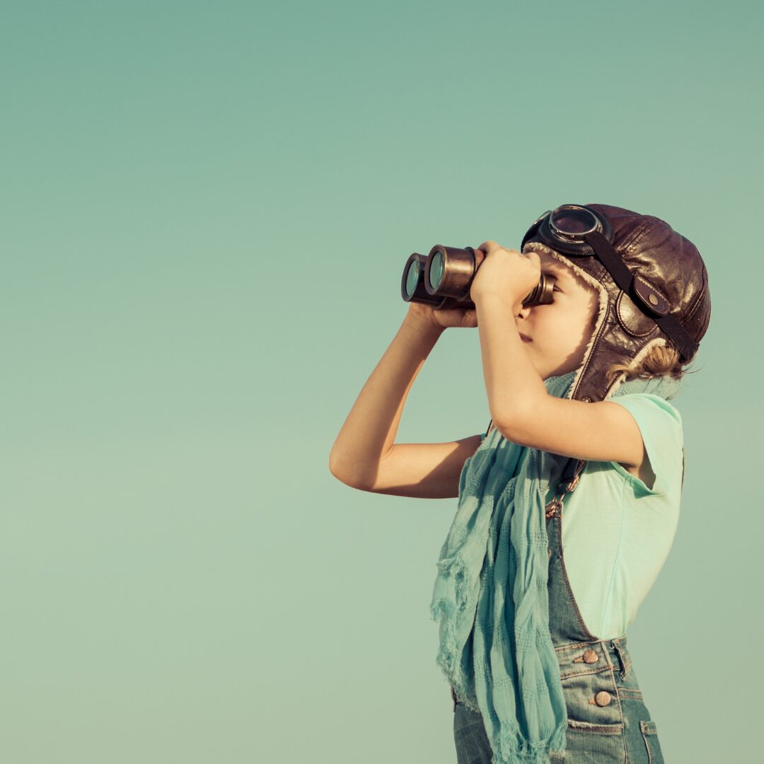 Una niña con gorro de aviador mira el horizonte con unos prismáticos, sobre un fondo azul verdoso
